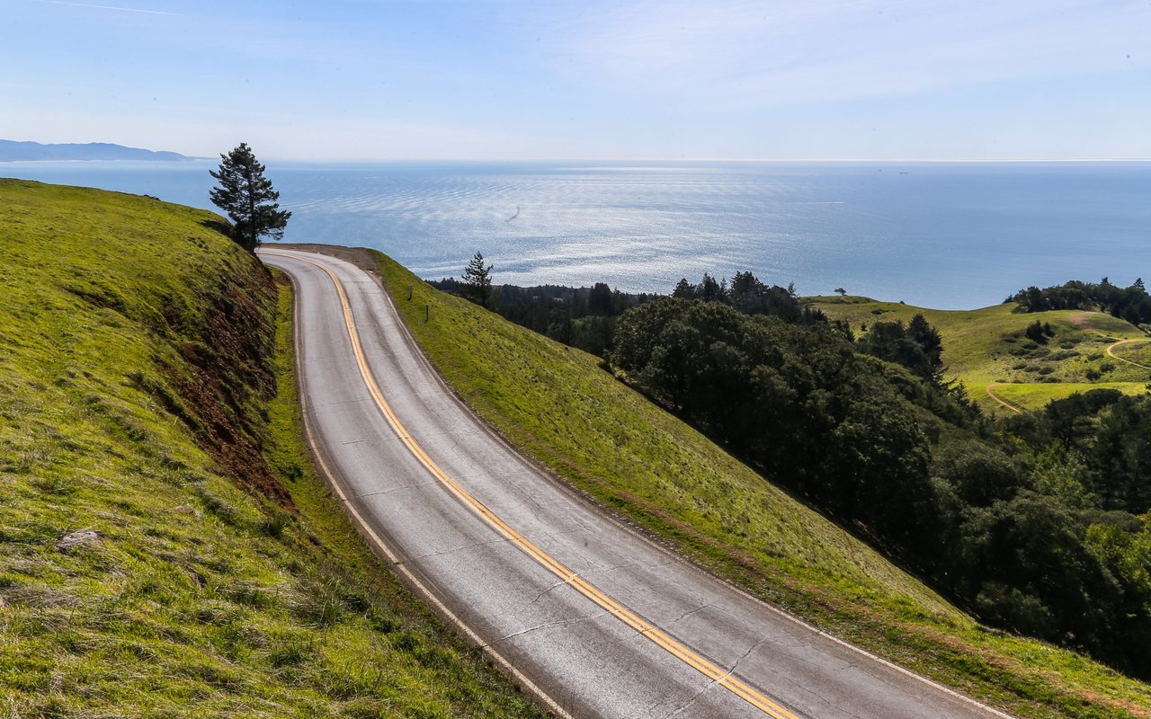 A winding coastal road crosses green hills above the ocean.