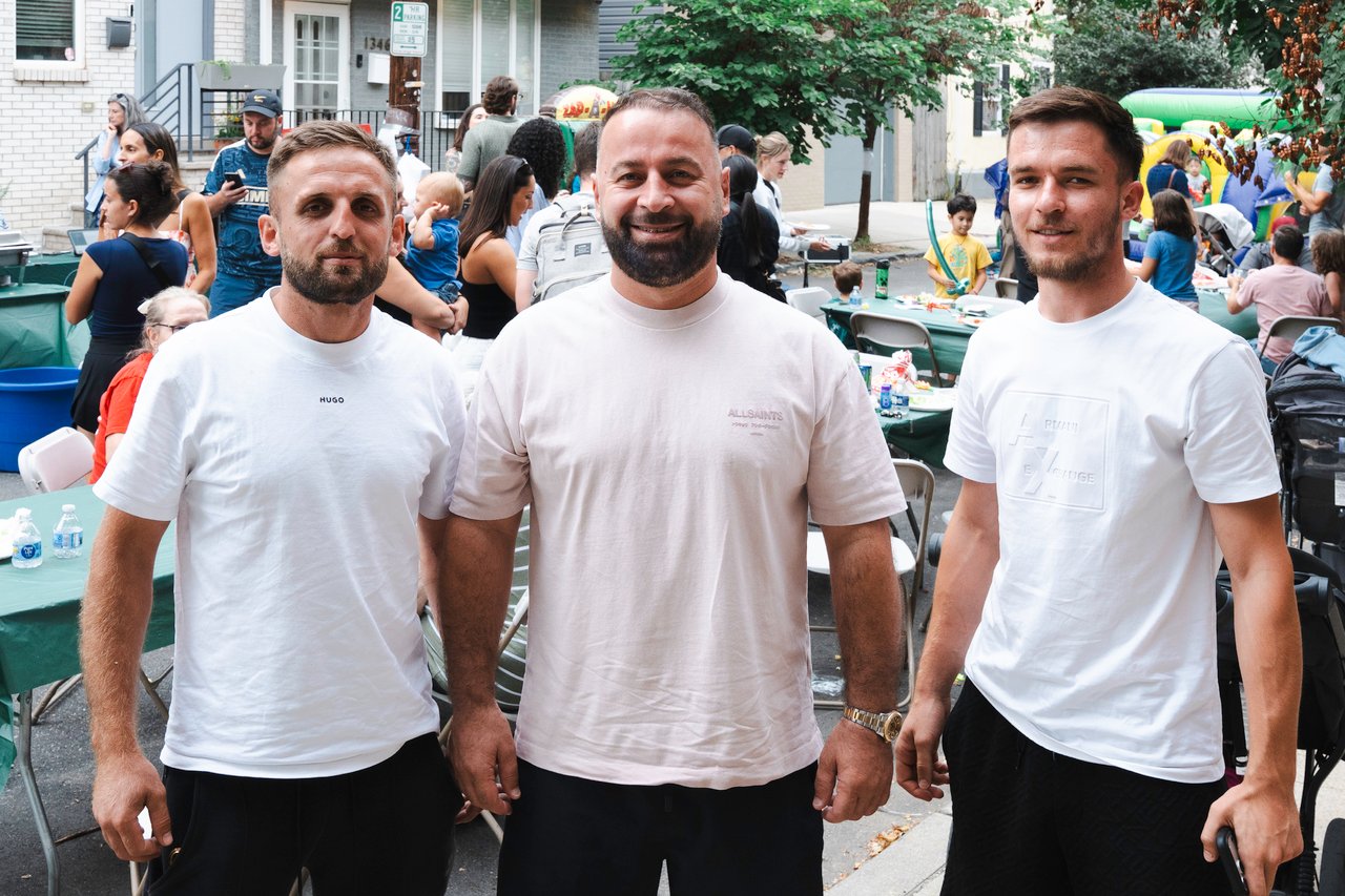 Three men pose at a neighborhood block party, with guests and tables behind them.