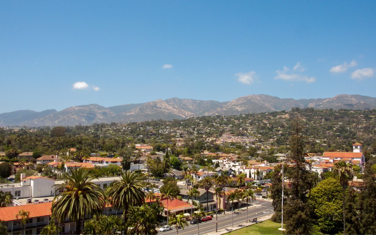 A scenic view of Santa Barbara with red-tiled rooftops, palm trees, and the Santa Ynez Mountains in the background.