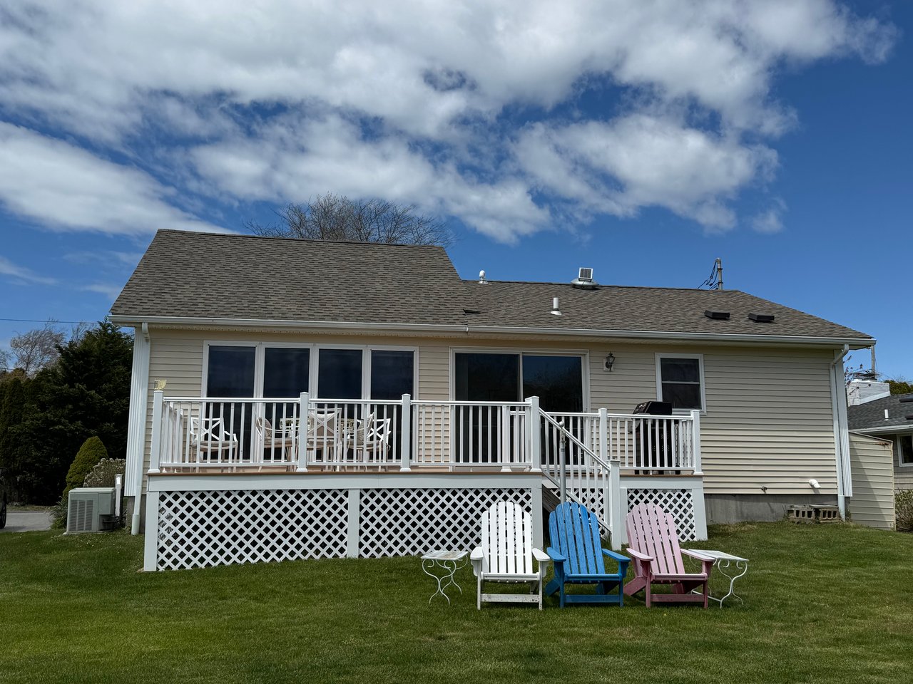Rear of a beige house with a raised white deck, outdoor dining set, and colorful Adirondack chairs on the lawn.