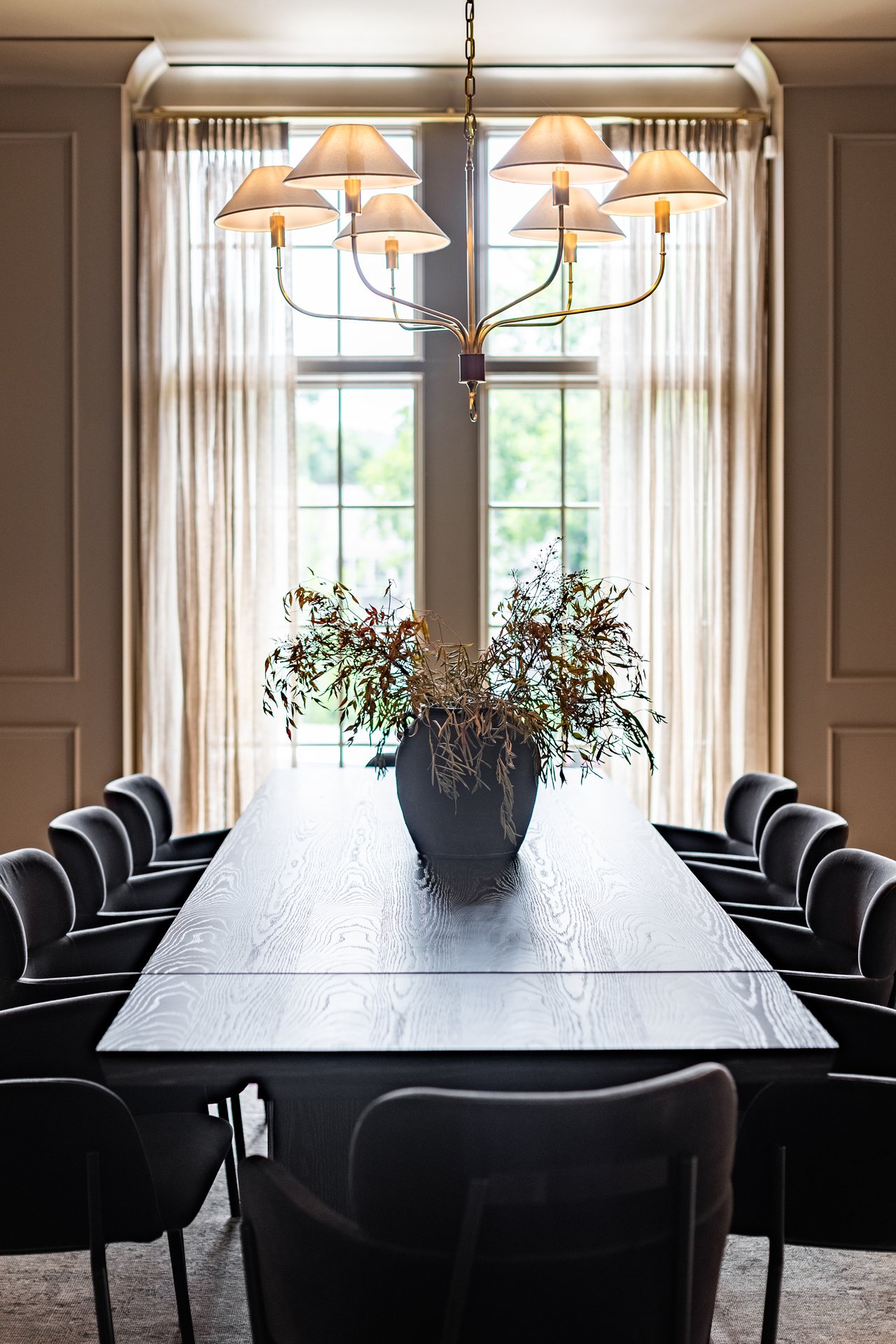 Formal dining room with a long pale wood table, dark chairs, and a chandelier beneath tall windows.