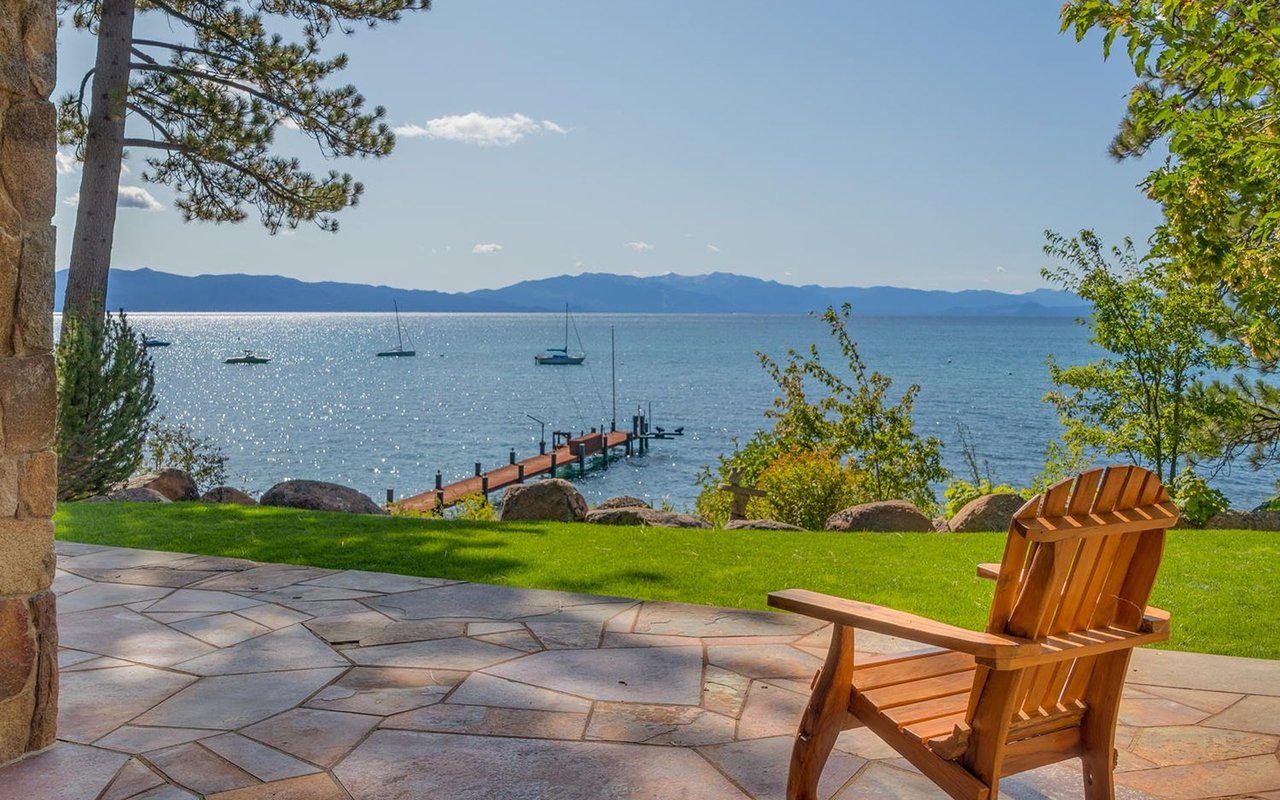Lakefront patio with a wooden Adirondack chair, private dock, sailboats, and distant mountains.