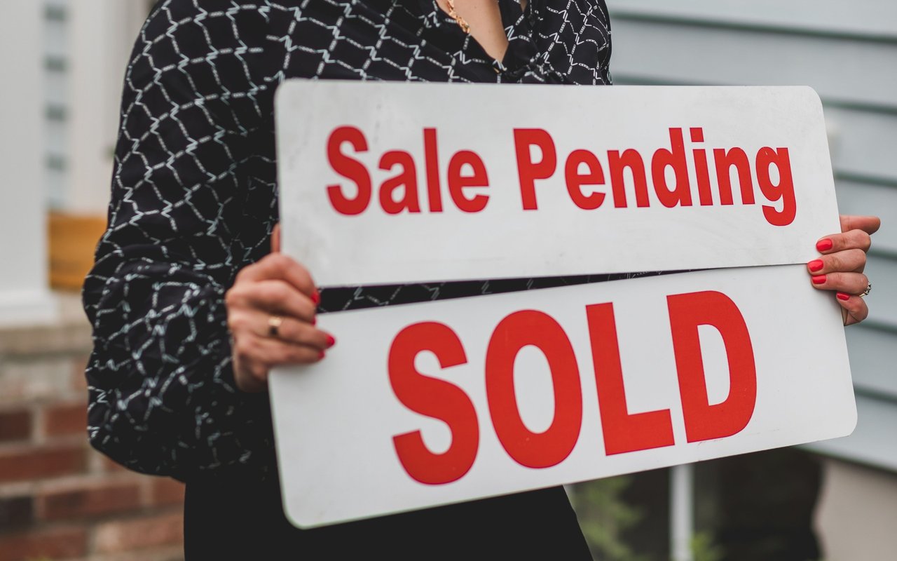 Person holding “Sale Pending” and “SOLD” signs outside a house