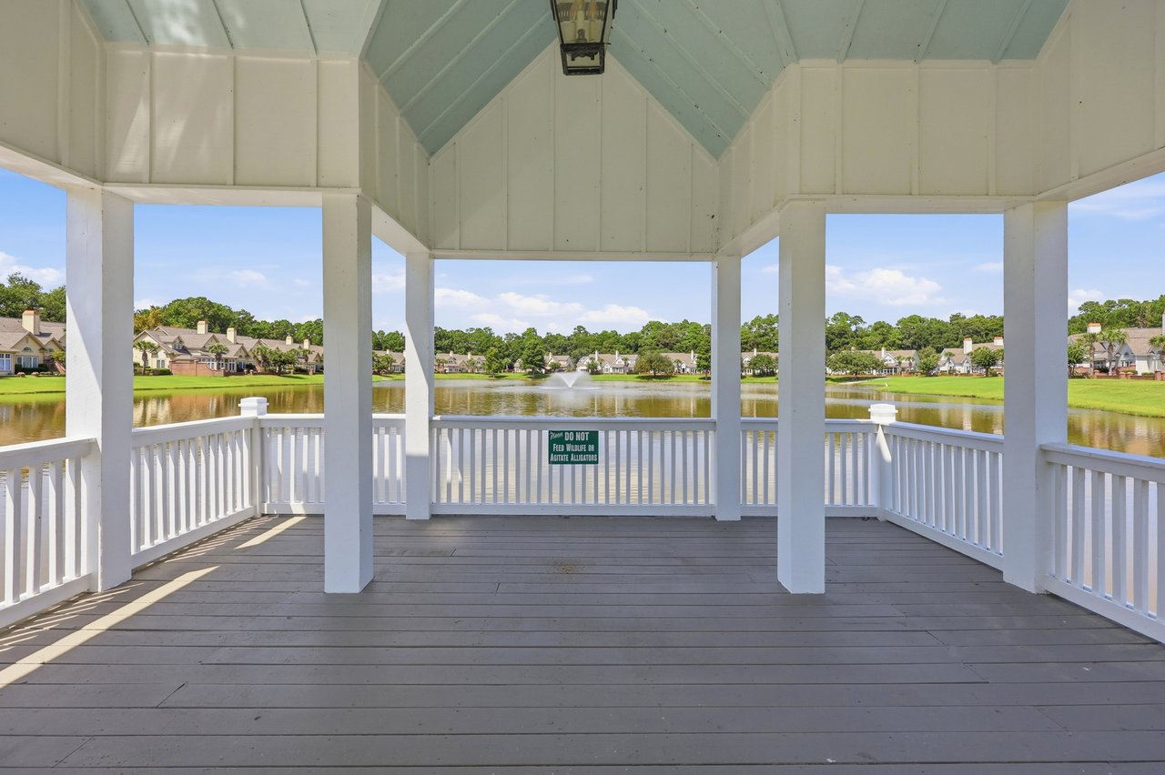Open-sided white gazebo overlooking a lake, fountain, and homes along the opposite shore.