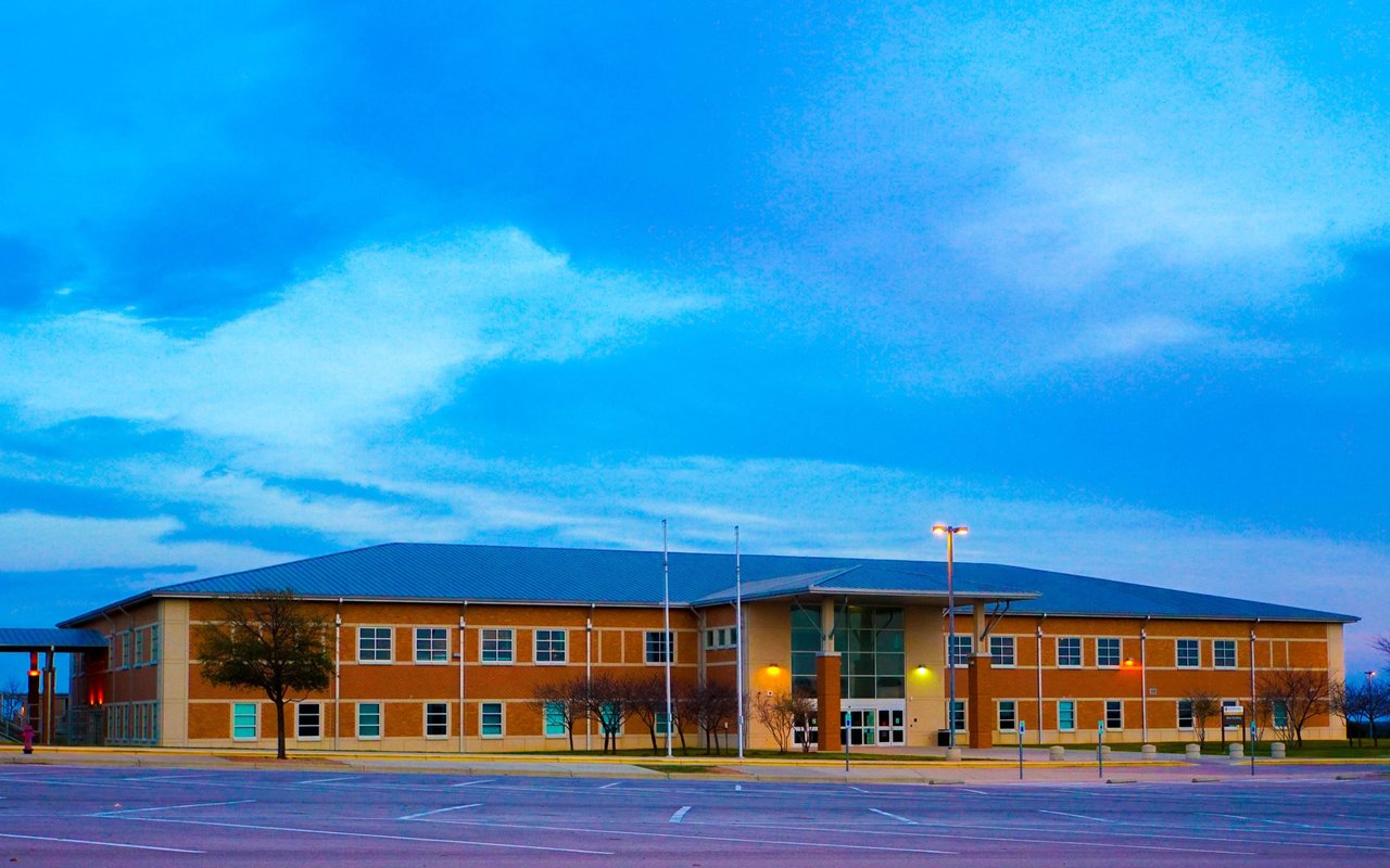 Two-story brick building with a blue roof and glass entrance beside an empty parking lot.