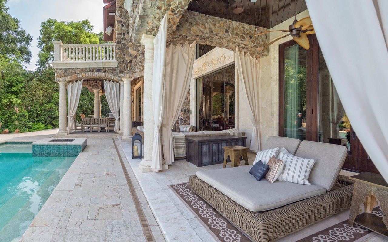 Stone patio with cushioned wicker seating beside a pool, framed by flowing curtains.
