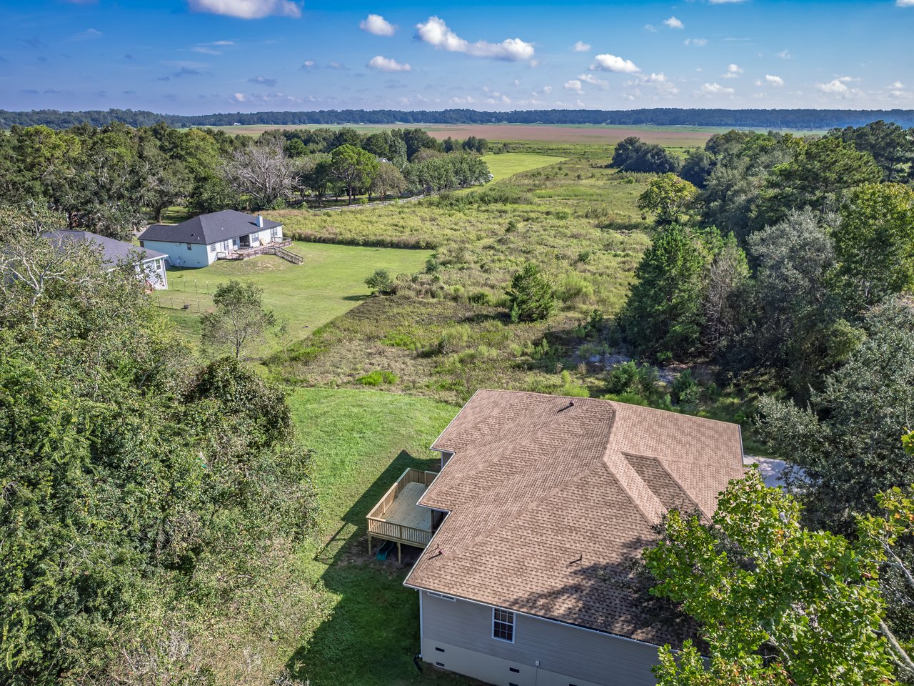 Aerial view of a home beside a broad lawn, trees, and open marshland.