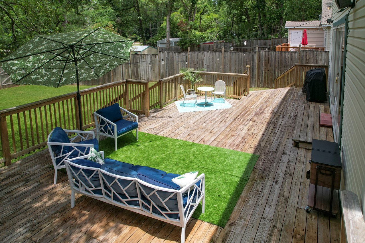 A wooden deck features blue cushioned seating, a patio umbrella, a small faux grass area, and a grill. It overlooks a green backyard with a fence.