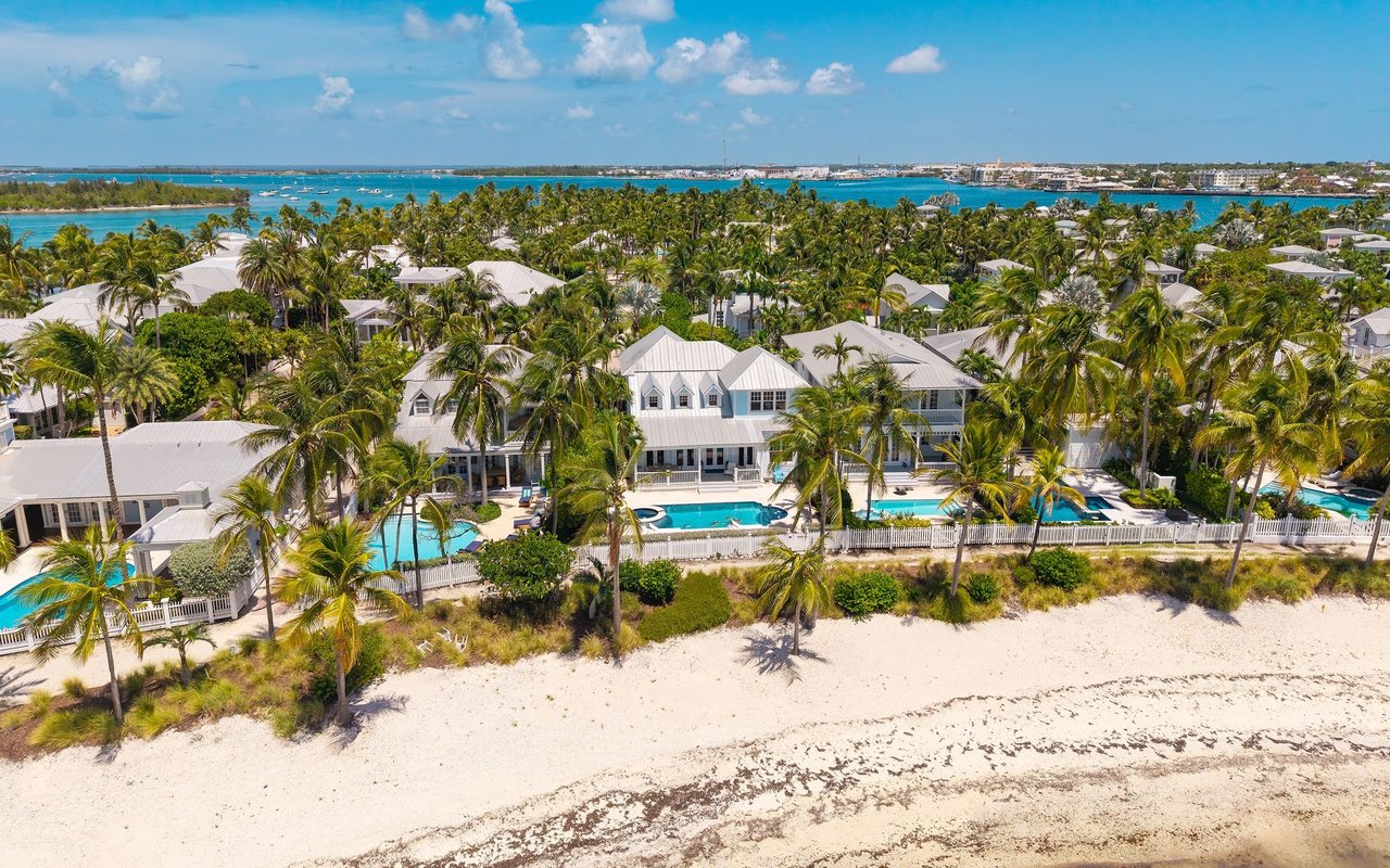 Beachfront homes with pools and palm trees beside a sandy beach and turquoise water.