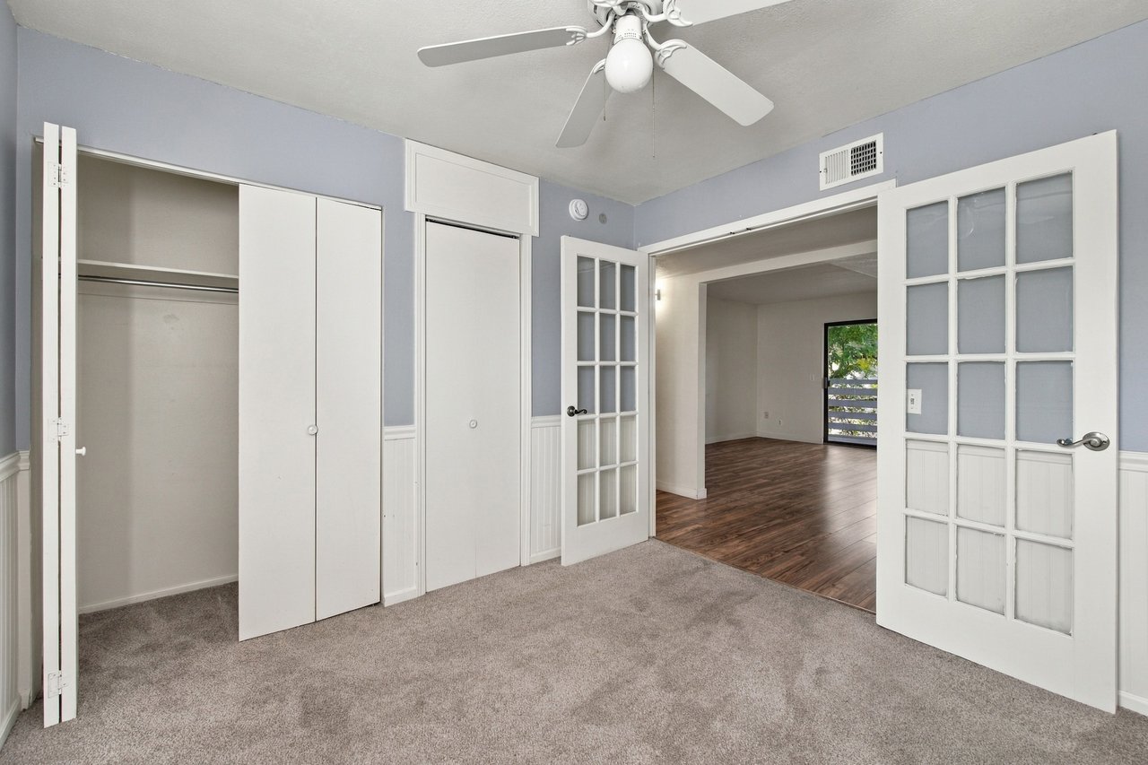 Empty bedroom with gray carpet, open closet, and French doors to a wood-floored room.