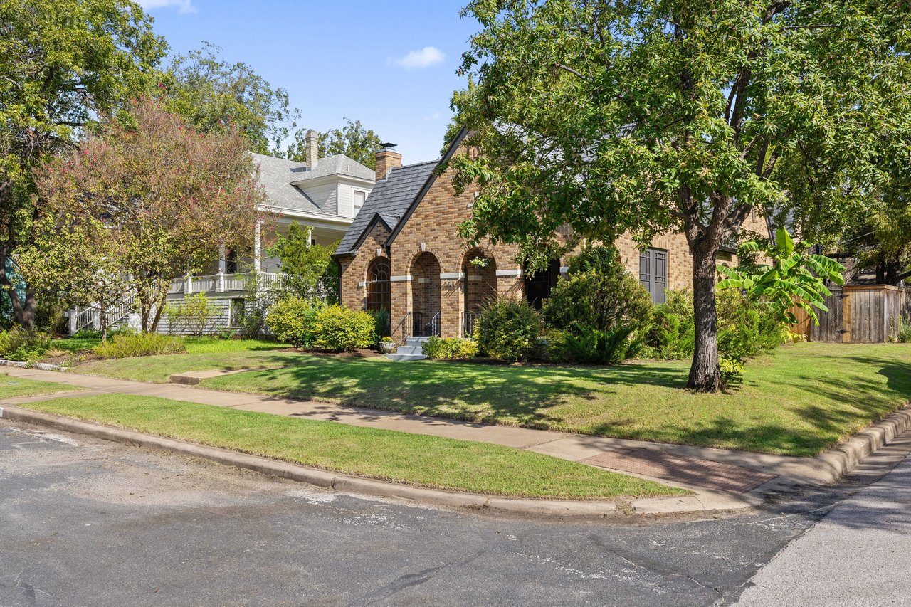Brick house with arched entry, steep roof, and mature trees along the lawn.