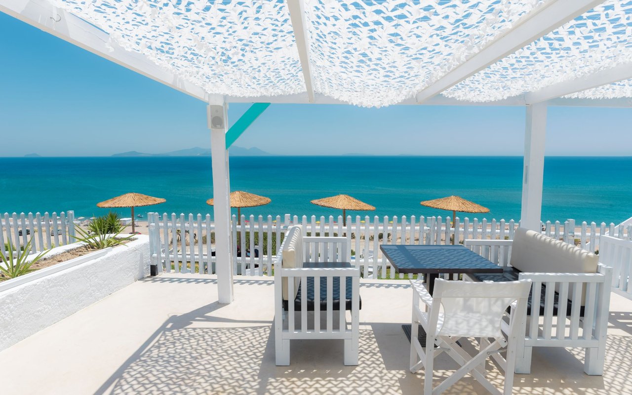 Seaside patio with white outdoor furniture beneath a patterned canopy, overlooking the beach and ocean.