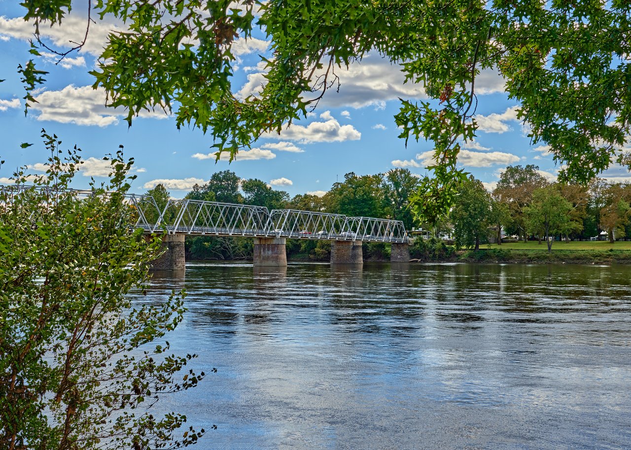 A metal pedestrian bridge crosses a broad river beneath leafy branches.
