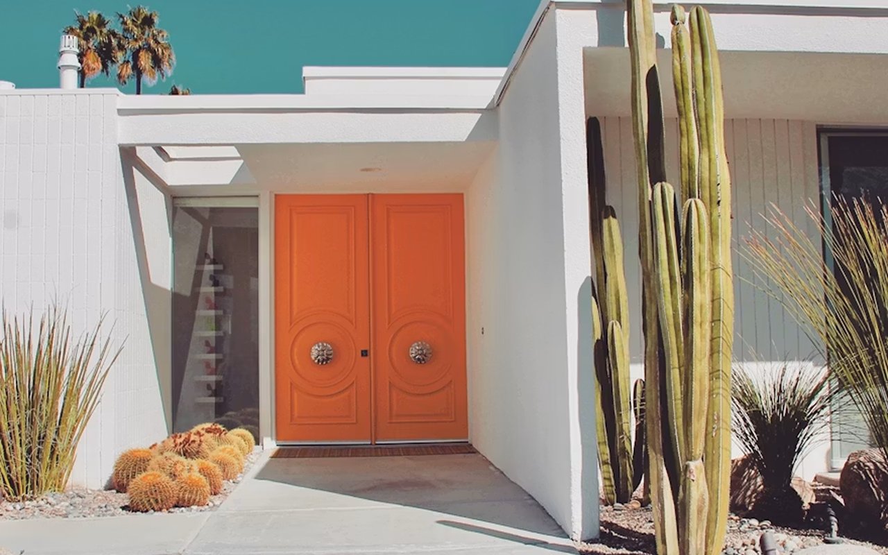 Modern white home entrance with orange double doors and tall cacti.