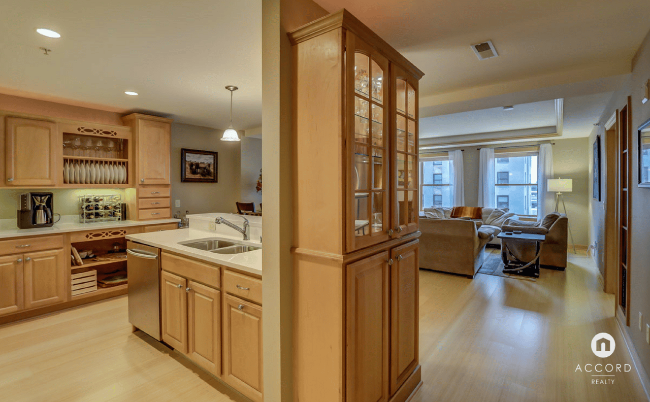 Condo kitchen with warm wood cabinetry opens onto a furnished living room with large windows.