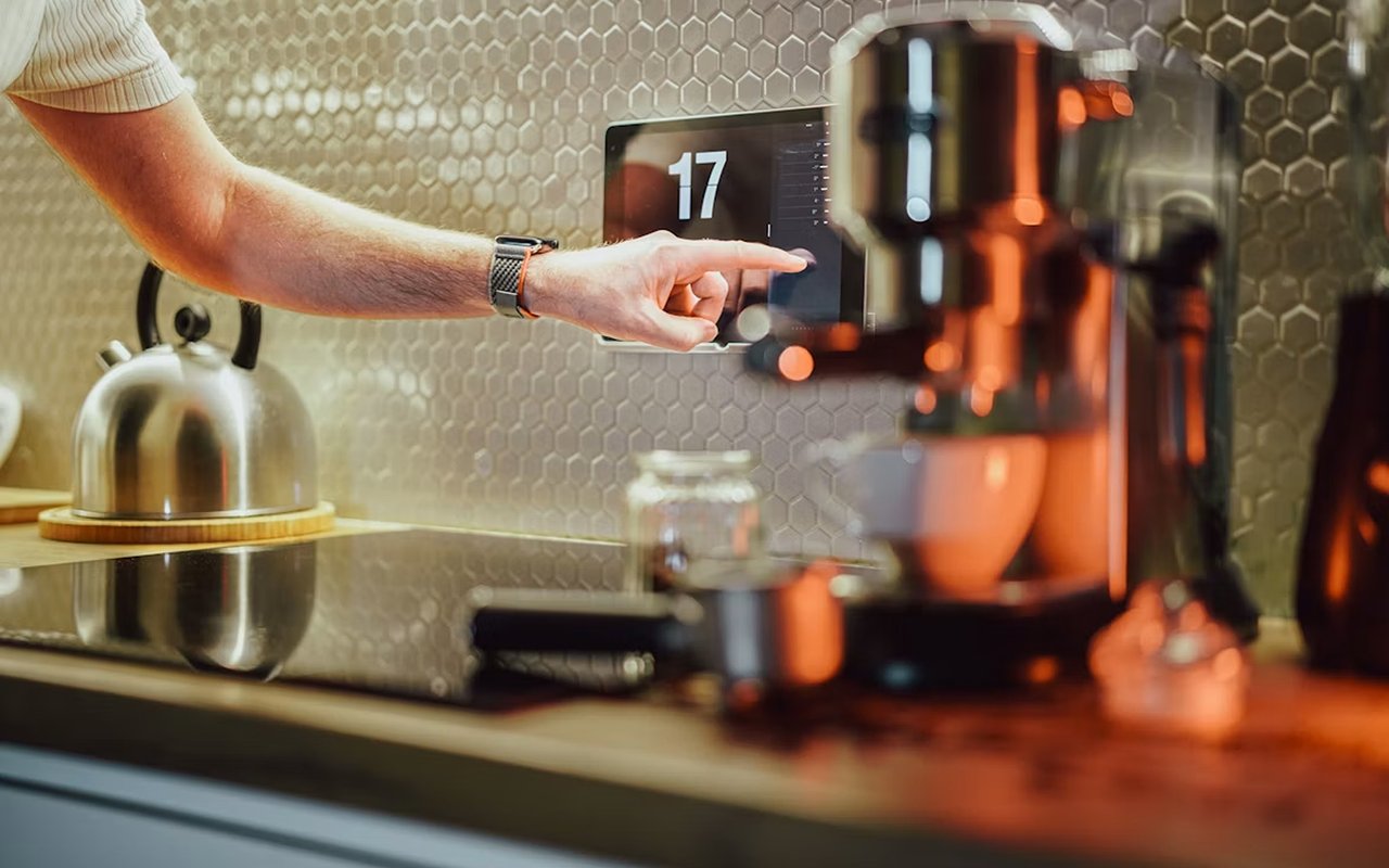 Modern kitchen counter with espresso machine, kettle, and a hand reaching toward a digital panel.