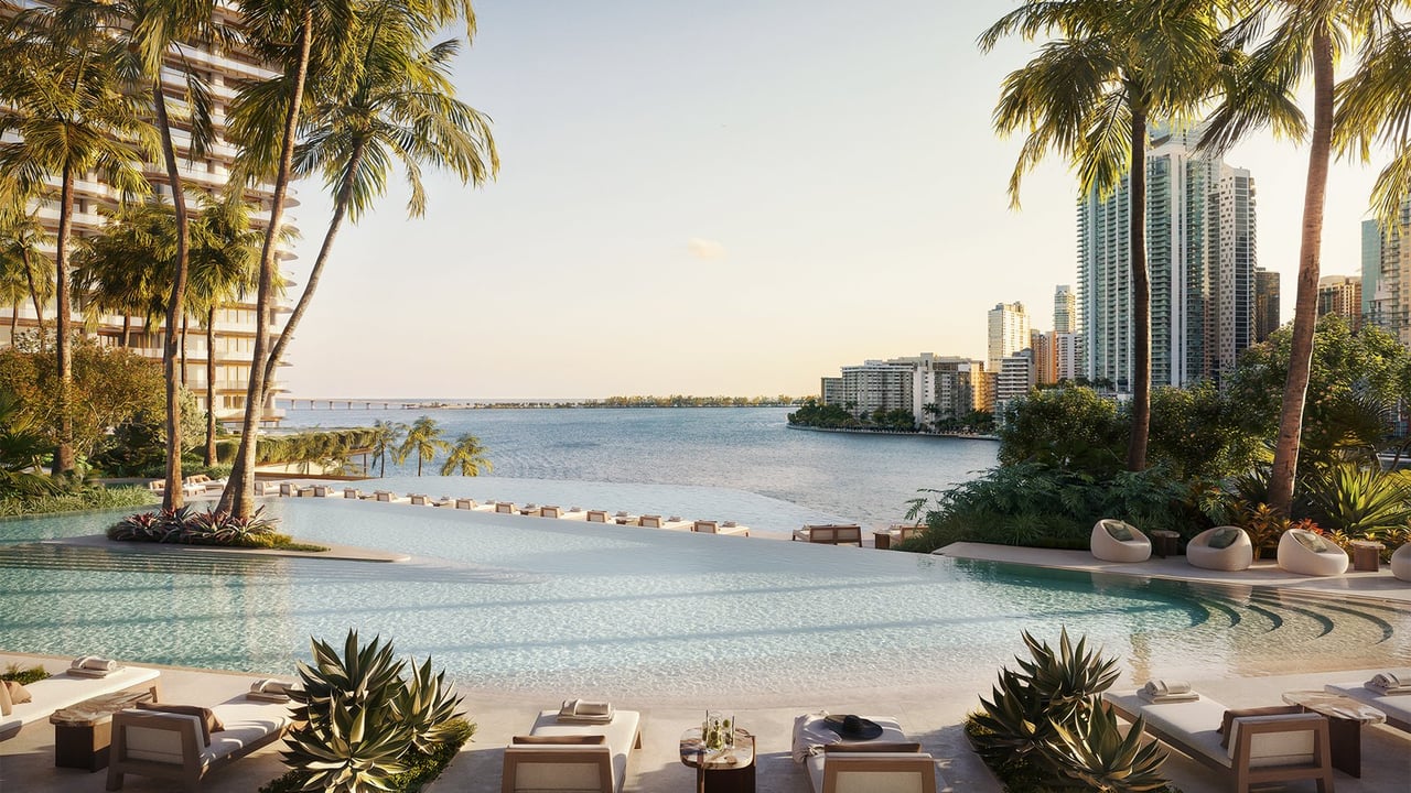 Waterfront pool terrace with palm trees, lounge chairs, and Miami high-rises across the water.