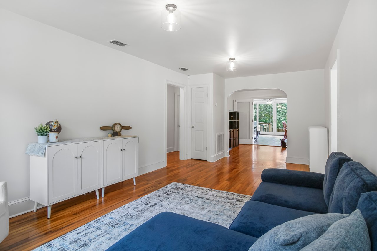 A bright living room with white walls and wood floors features a blue sofa, white sideboard with decor, and arched doorway leading to a sunlit room.
