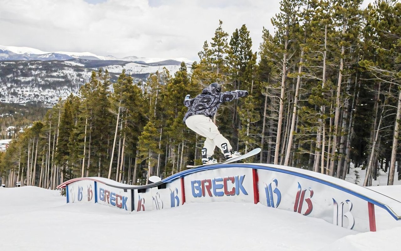 Snowboarder jumping a BRECK rail in a snowy terrain park, with forested mountain ridges behind.