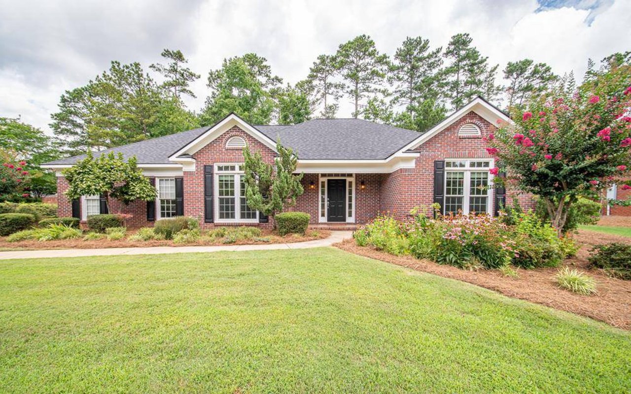 Red brick home with a gray gabled roof, black shutters, and flowering landscaping across the front yard.