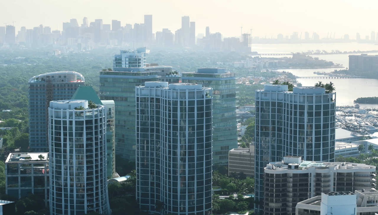 Waterfront condominium towers rise above Coconut Grove greenery, with the bay, marina, and distant skyline beyond.