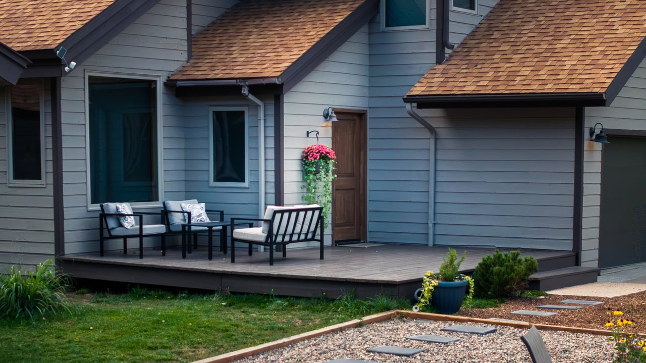 Gray-sided home with a covered porch, outdoor seating, hanging flowers, and a gravel garden path.