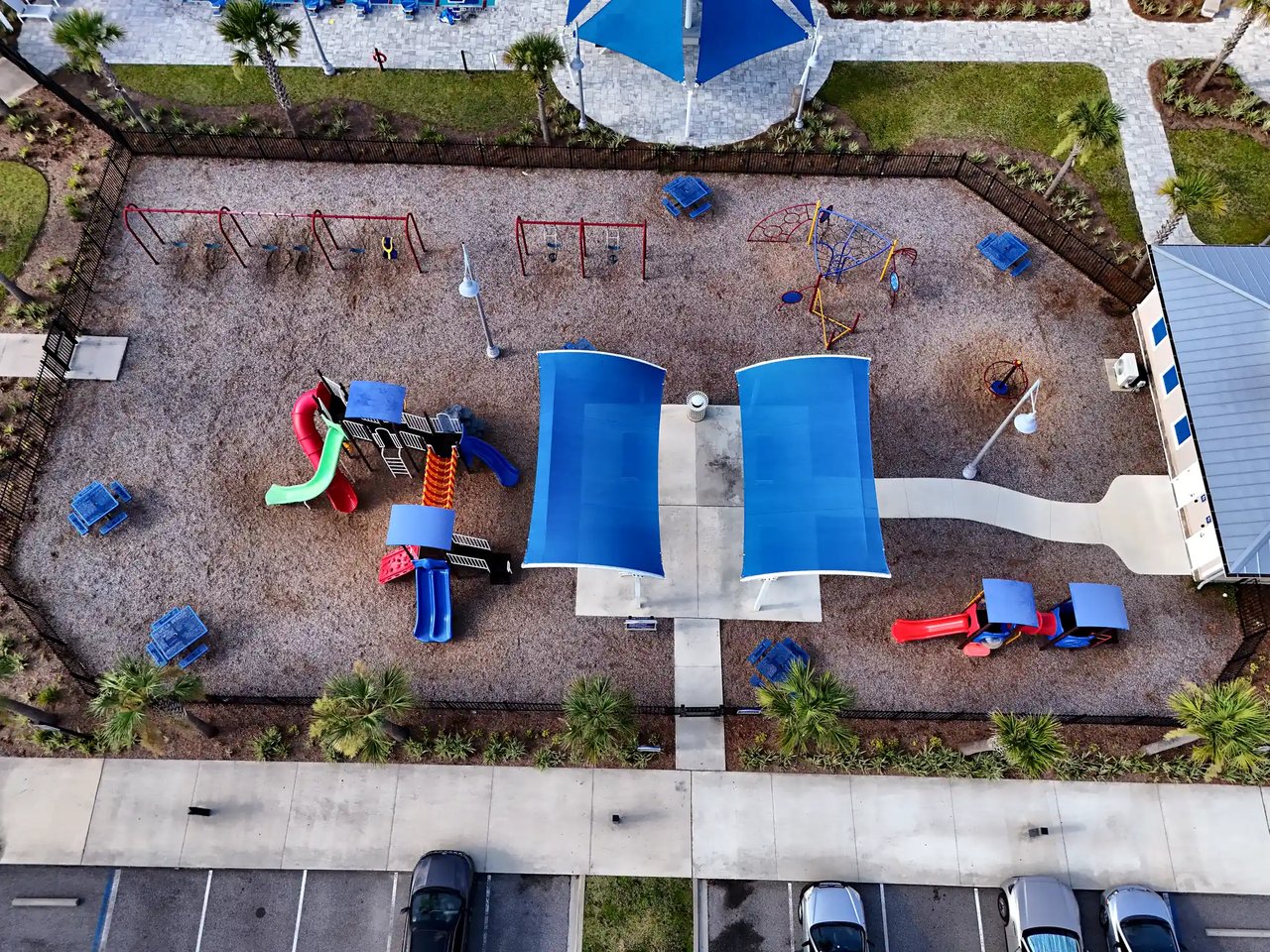 Fenced community playground with colorful play equipment and blue shade canopies, seen from above.