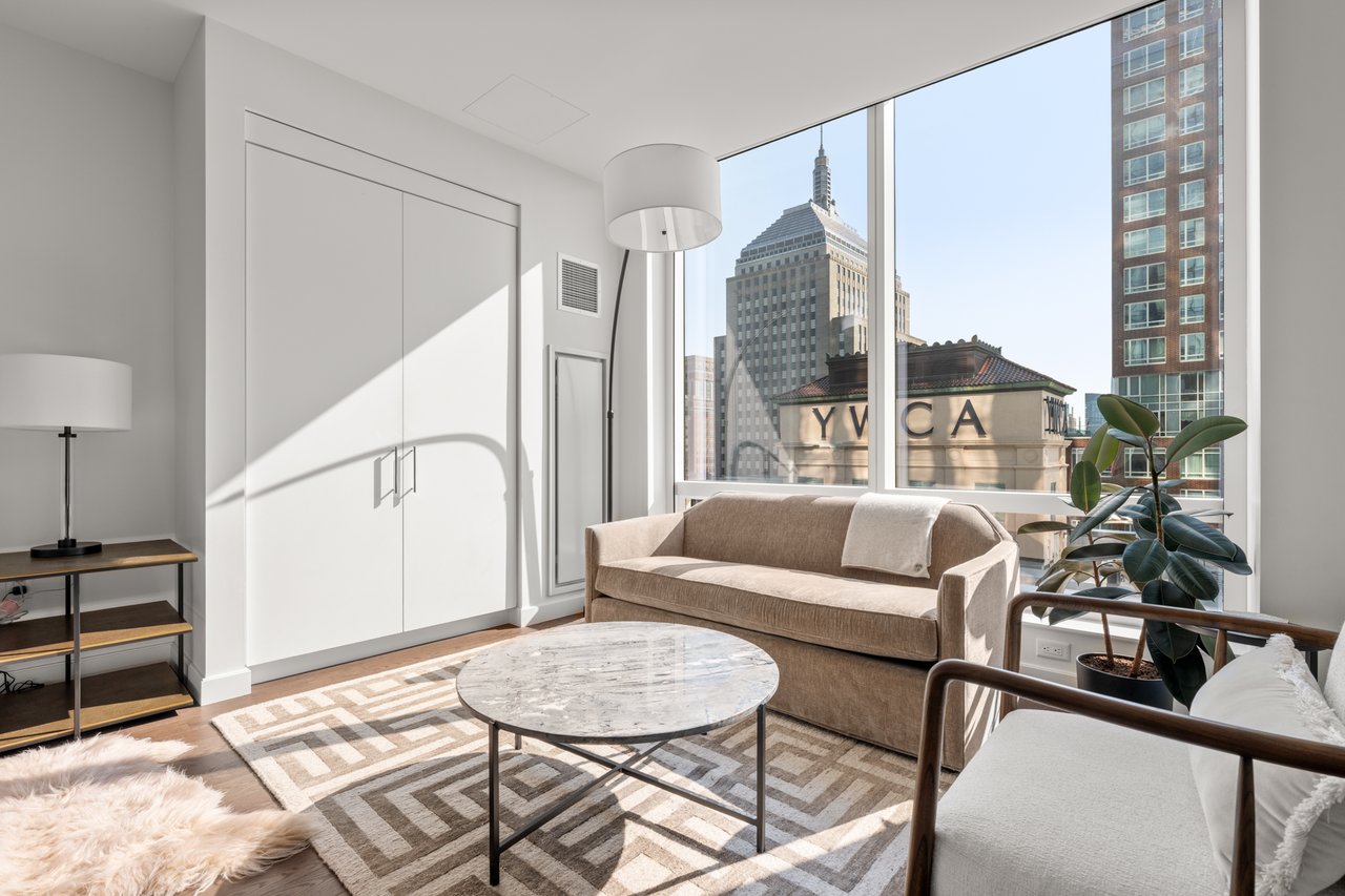 Living room with floor-to-ceiling windows overlooking city buildings, a beige sofa, and a marble coffee table.