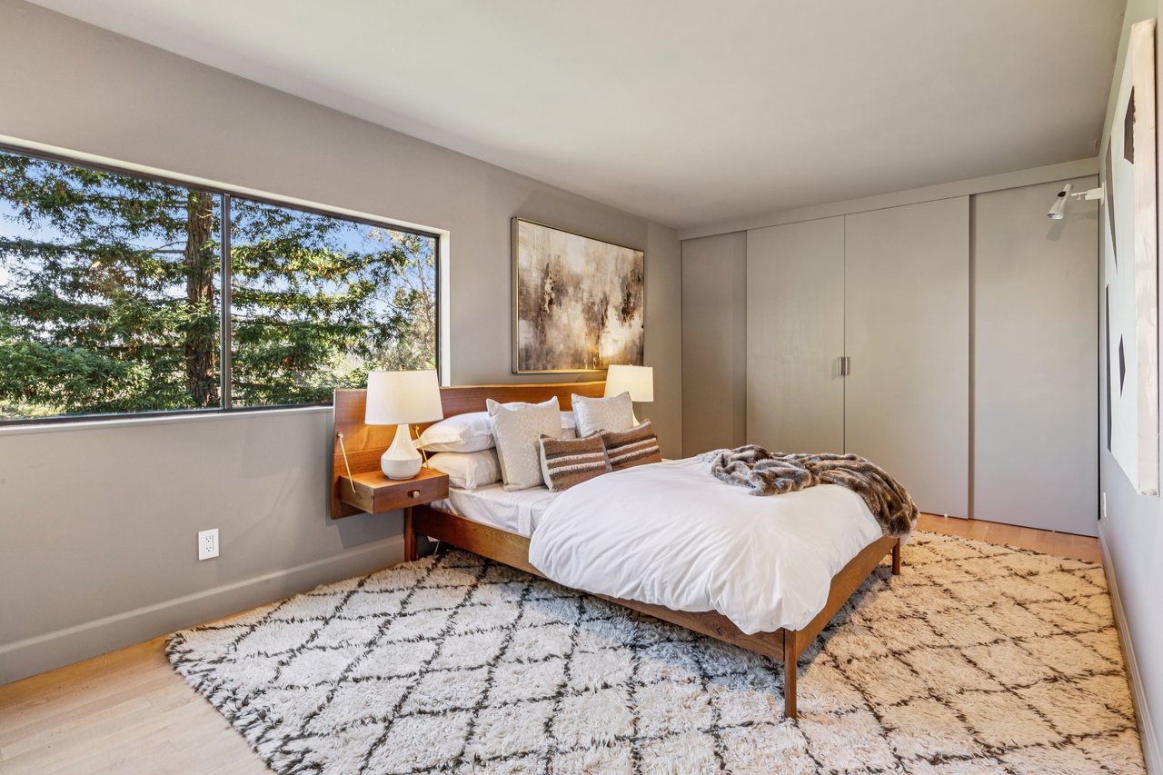Bedroom with a wood platform bed and a large window overlooking leafy trees.