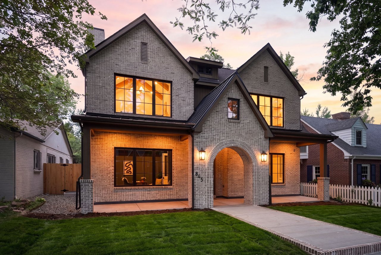Two-story brick house with a gray arched entry and warmly lit windows at dusk.
