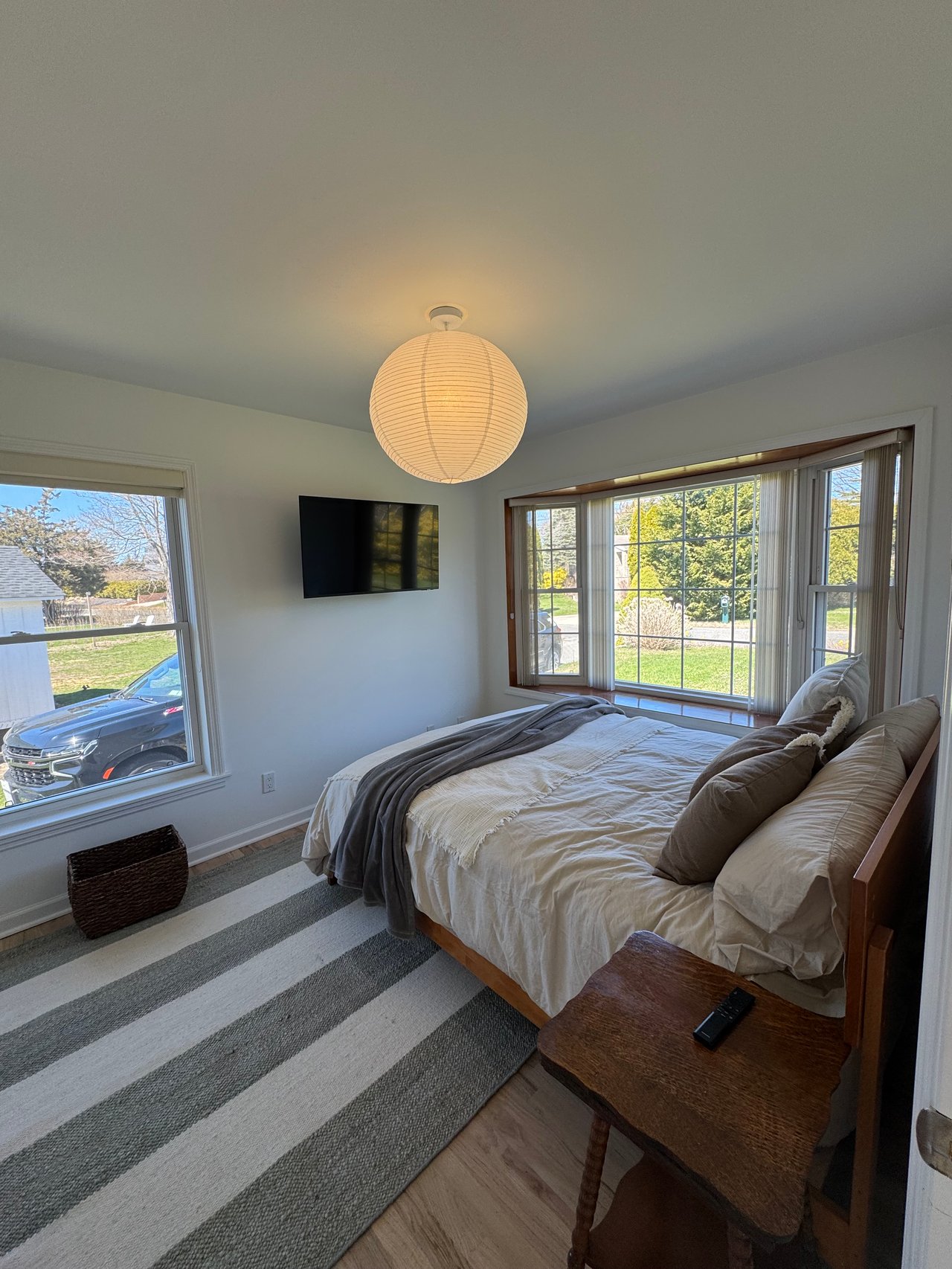 Furnished bedroom with a striped rug, round paper lantern, and large windows overlooking the yard.
