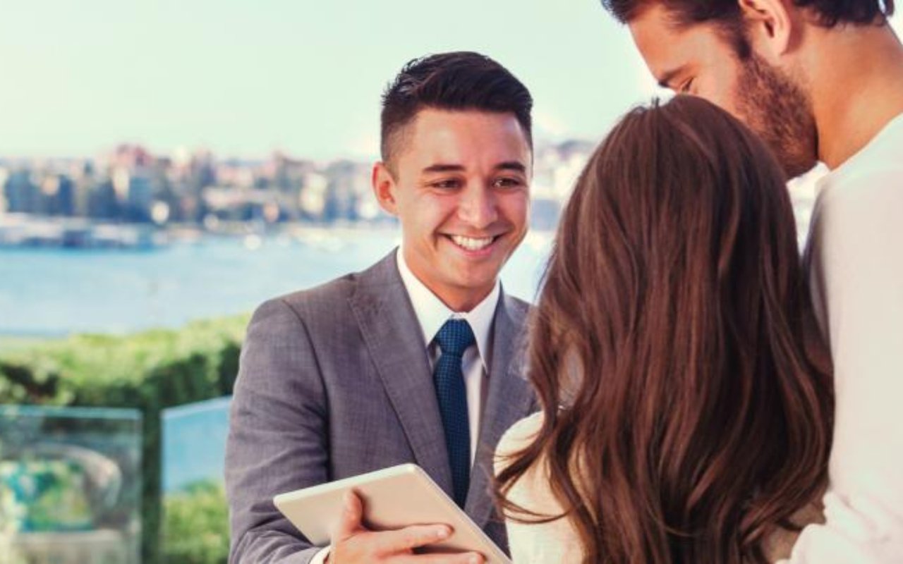 A suited real estate agent shows a tablet to a couple beside a waterfront.