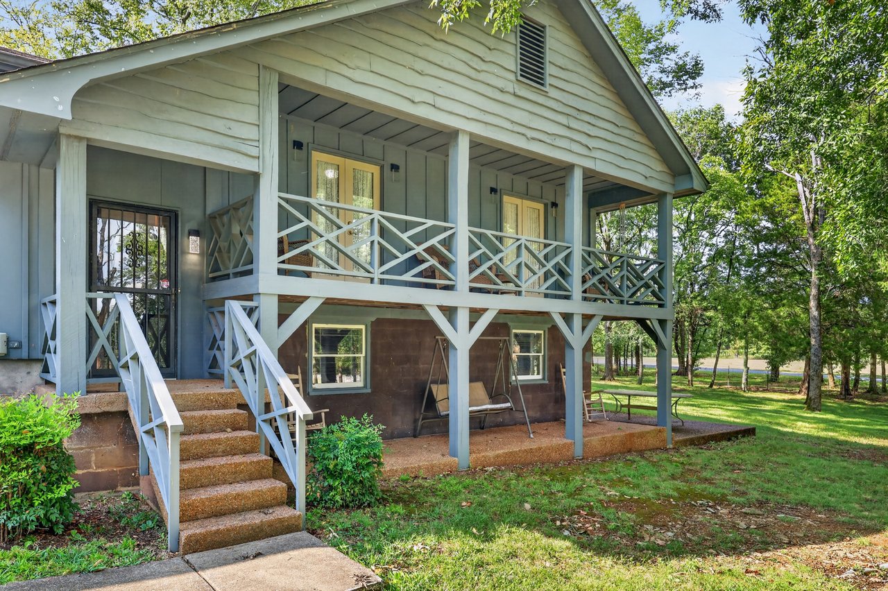 Raised house with a covered porch, cross-braced railings, front steps, and a tree-shaded lawn.