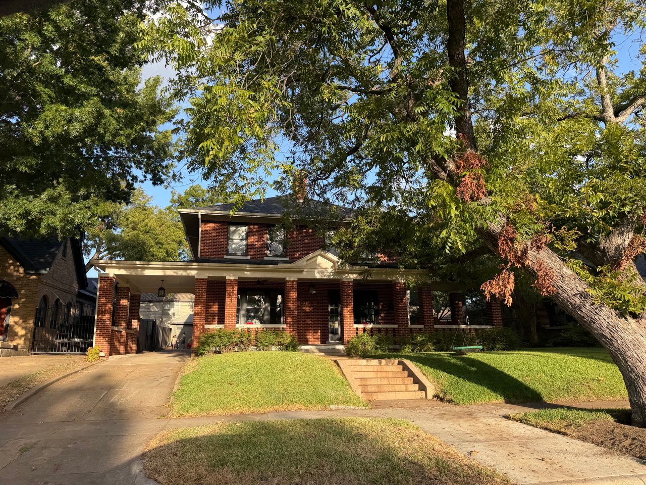 Two-story brick home with a covered porch beneath mature shade trees.