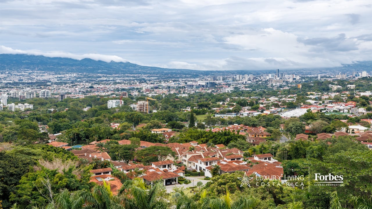 Panoramic Escazú view with red-tile homes, green canopy, city skyline, and cloud-covered mountains.