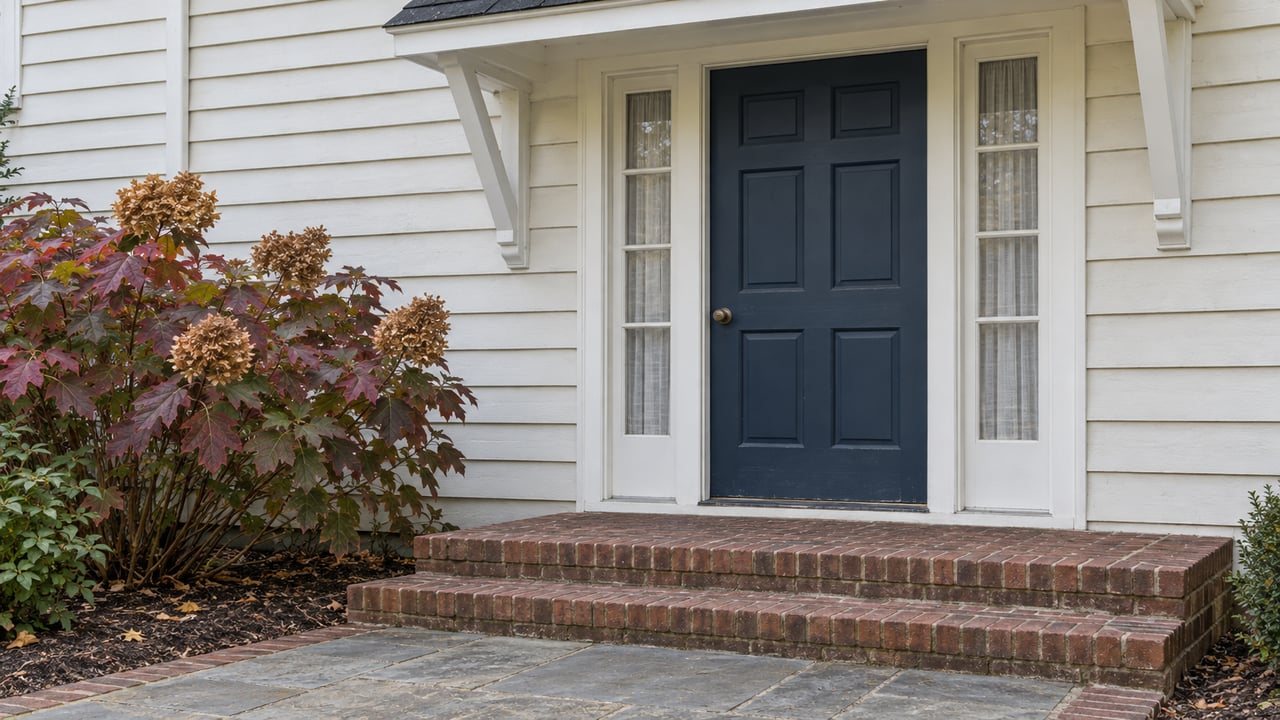 Close view of an ivory clapboard home with a dark blue door, paired sidelights, weathered brick steps and burgundy oakleaf hydrangea.