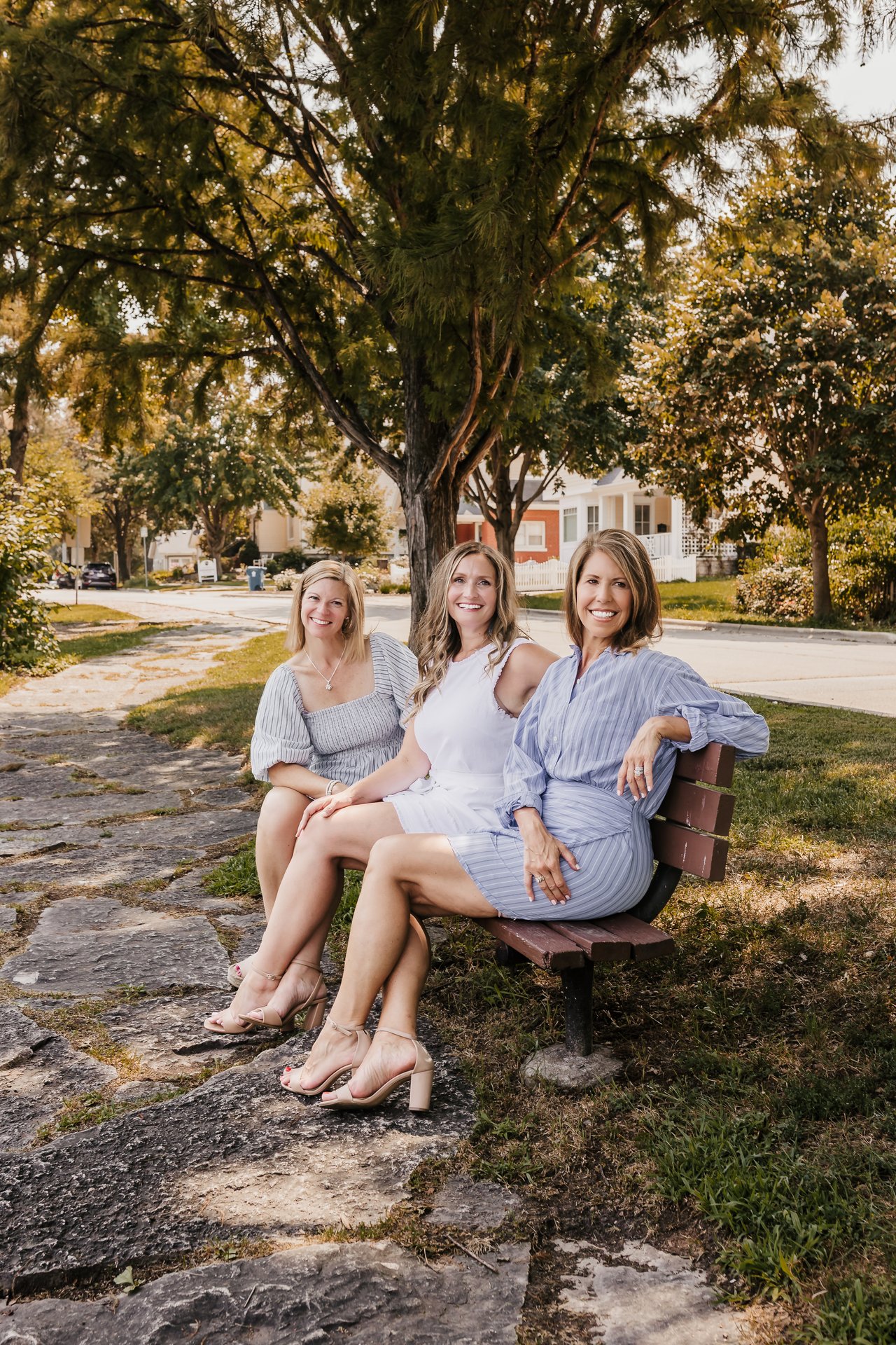 Three women sit smiling on a bench beneath a large tree on a residential street.