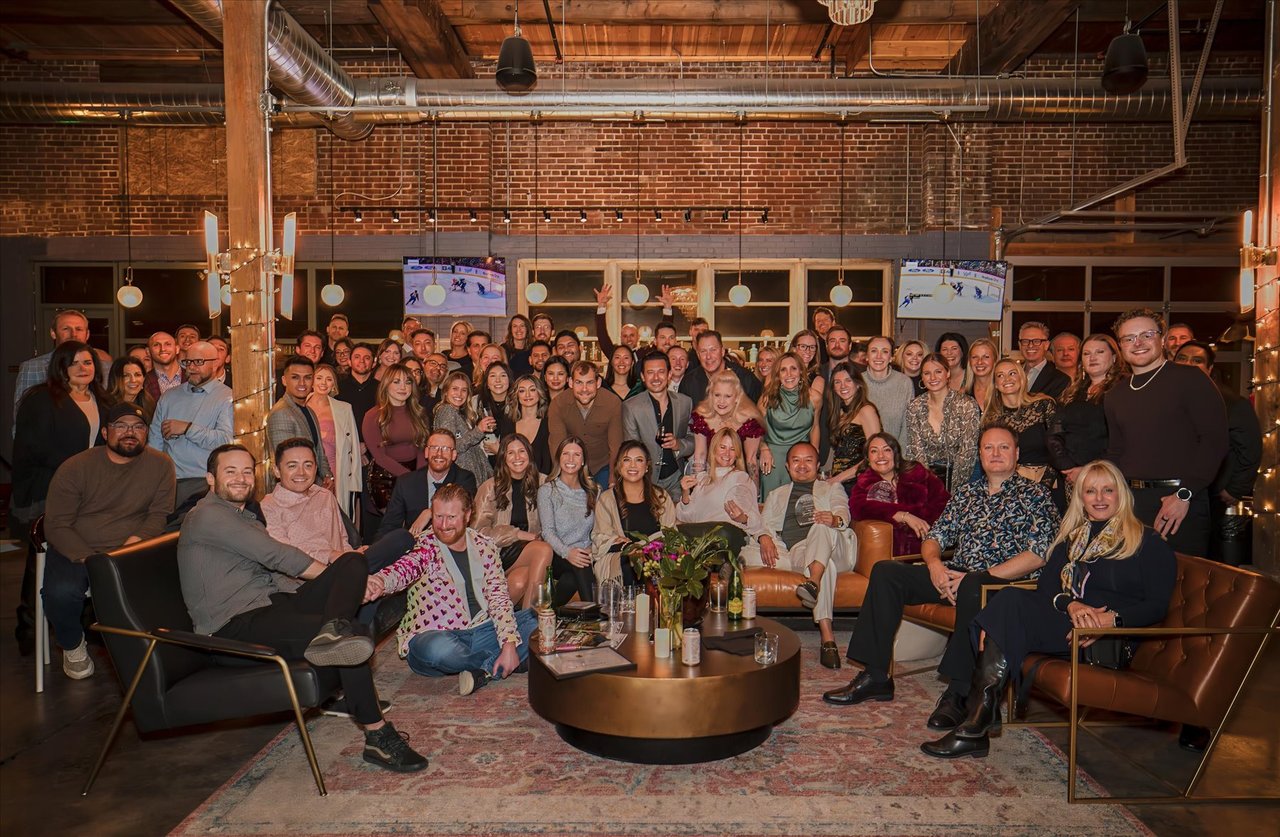 A large group poses in a brick-walled lounge with leather seating and exposed wooden beams.