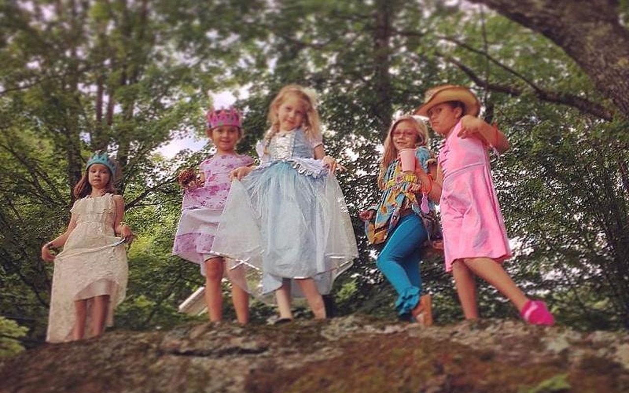 Children in colorful costumes pose together on a rocky ledge beneath leafy trees.