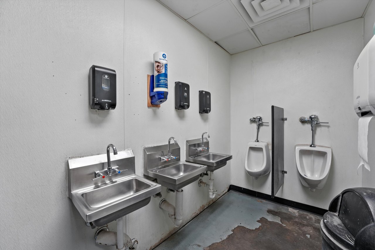Commercial restroom with three stainless-steel sinks, two urinals, and a worn concrete floor.