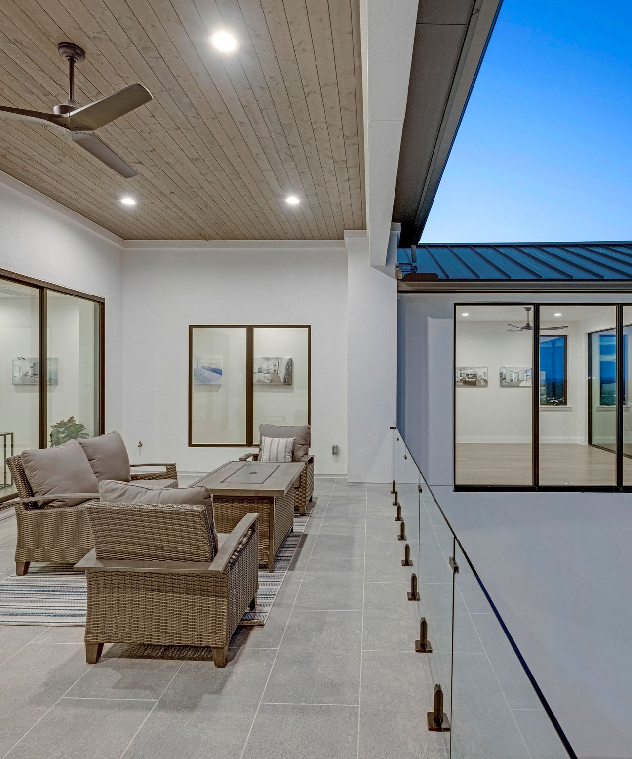 Covered patio with wicker seating, a wood-plank ceiling, and glass railing beneath a blue sky.