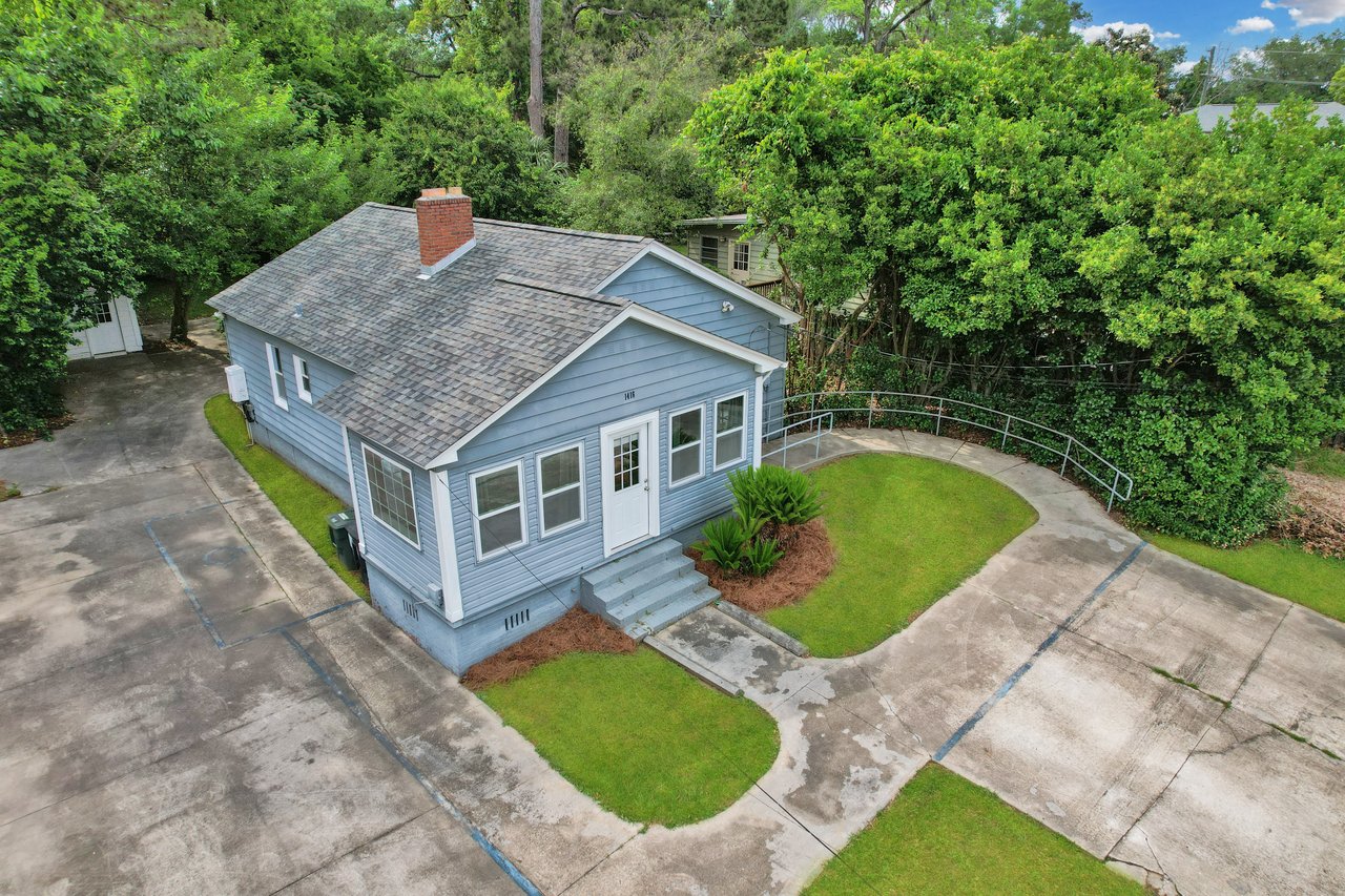 A small blue house with a gray shingle roof and brick chimney. It features a white door, surrounded by lush green trees and a driveway. Calm atmosphere.