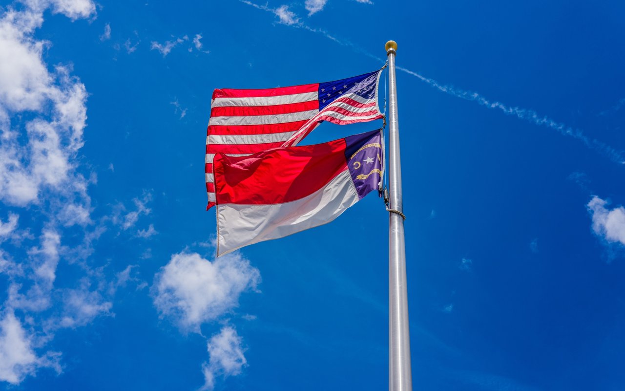 United States and North Carolina flags flying against a blue, cloud-dotted sky.
