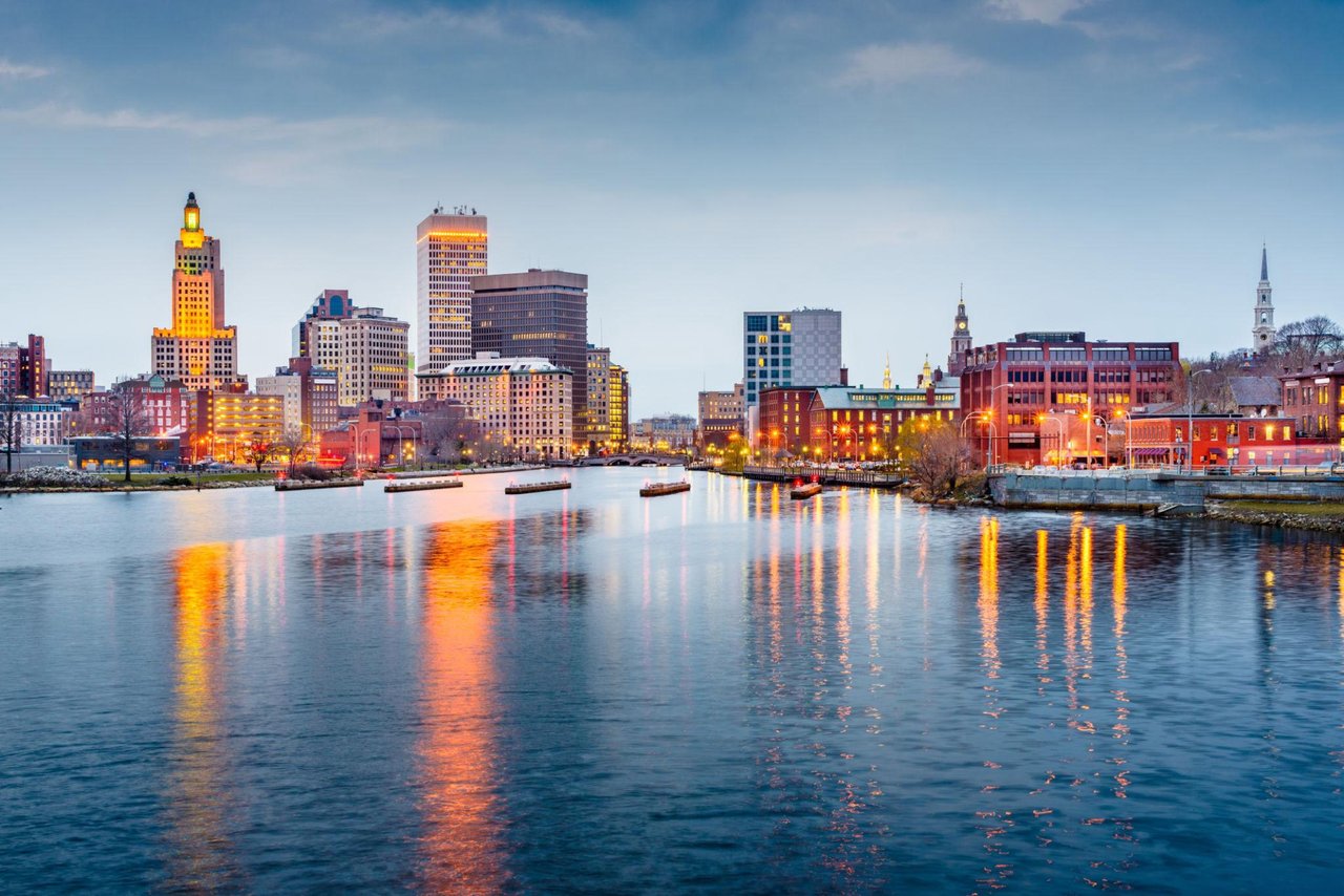 Providence skyline at dusk, with illuminated buildings reflected in the river.