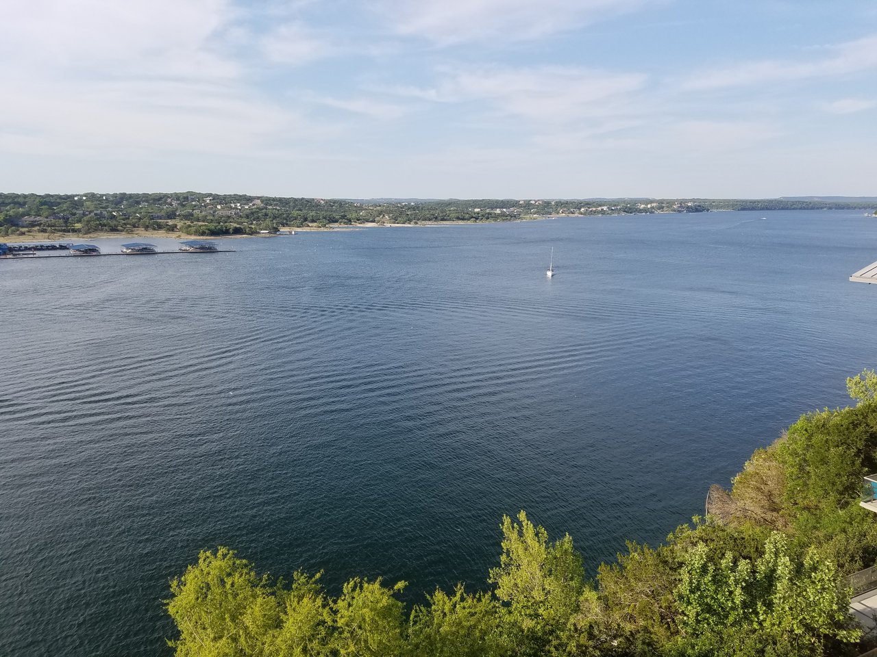 Blue lake with a distant tree-lined shore, a small sailboat, and leafy trees in the foreground.