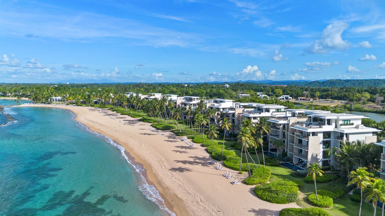 Aerial view of beachfront residences lining a sandy beach beside turquoise water and palm trees.