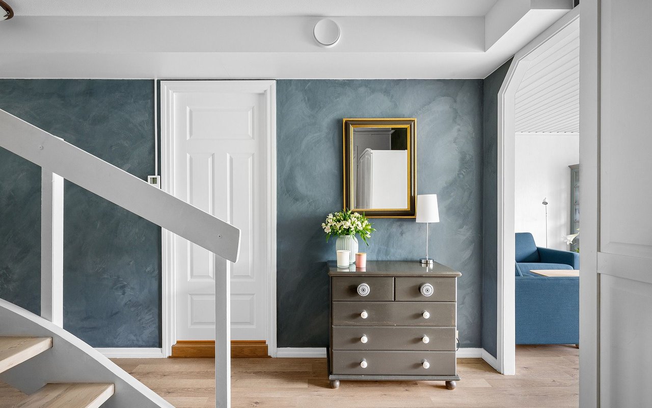Entry hall with blue walls, a white staircase, and a chest of drawers beneath a gold-framed mirror.