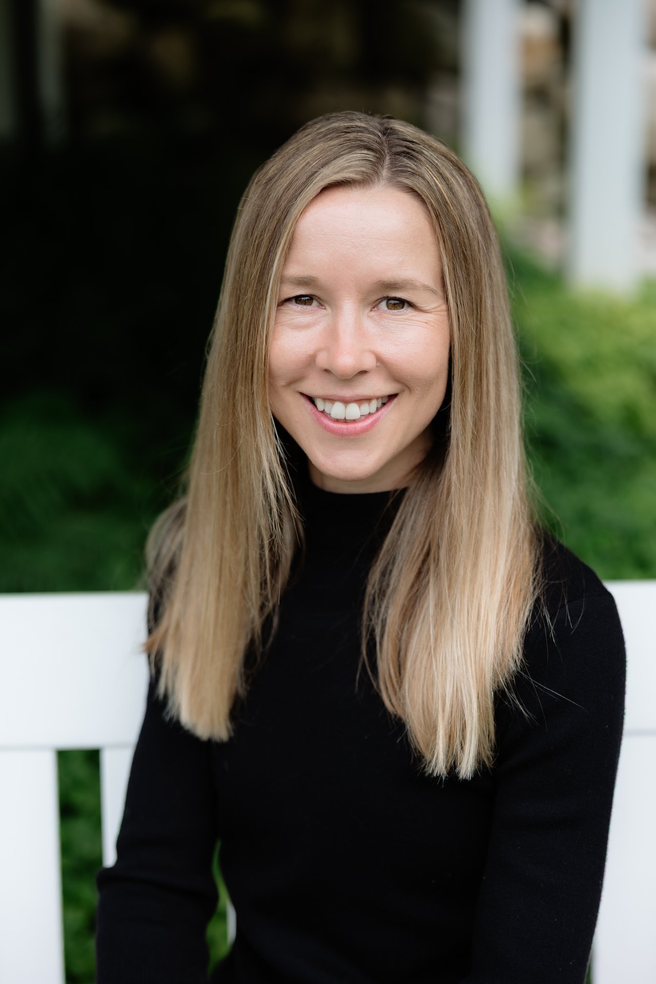 Dani Davis smiles in a black top, seated before a white bench and greenery.