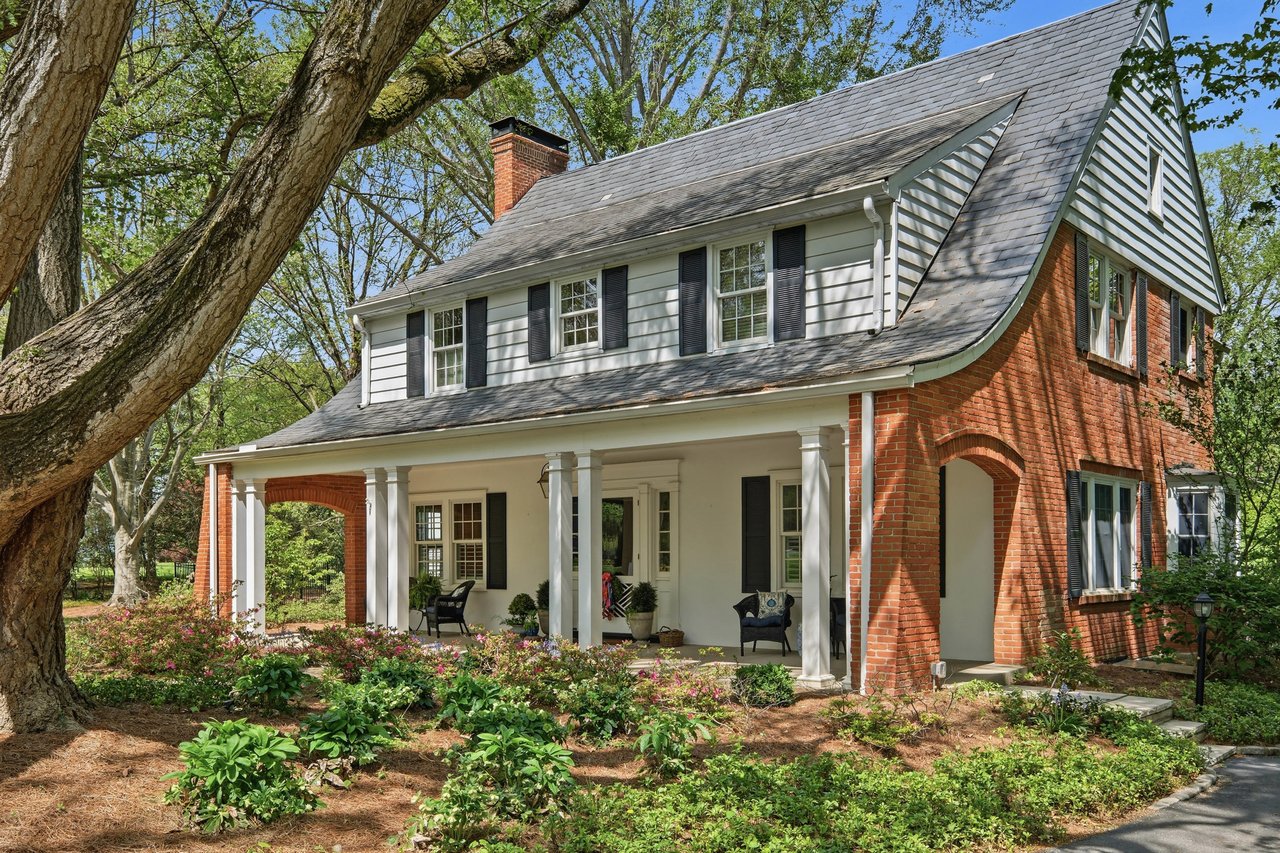 Red-brick and white-sided house with a columned porch, mature trees, and landscaped gardens.