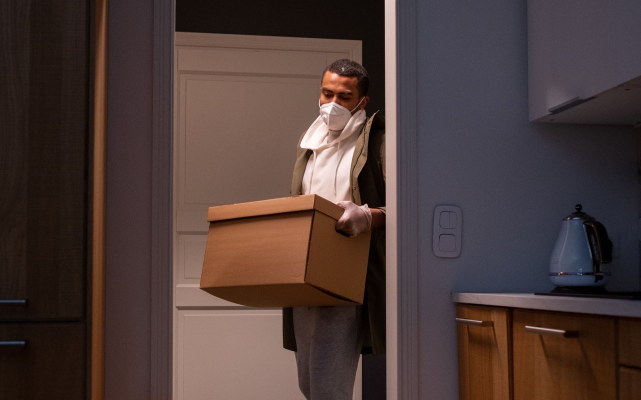 Masked person carrying a moving box through a home's doorway.